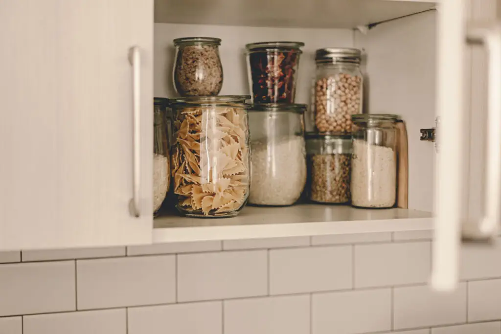 Pantry Organization: Glass jars filled with pasta, grains, and legumes neatly arranged on a shelf inside a white kitchen cabinet.