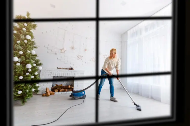 View through a window pane of a woman vacuuming a bright living room decorated with a Christmas tree, illustrating professional residential cleaning services preparing a home for the holidays.