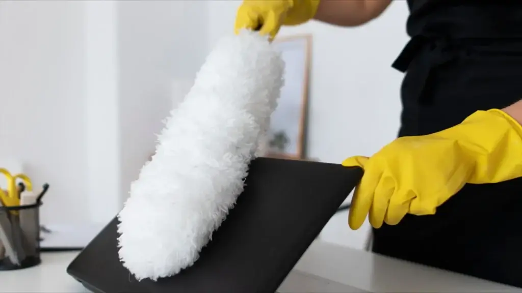 Hands in yellow gloves dusting a laptop with a white duster.