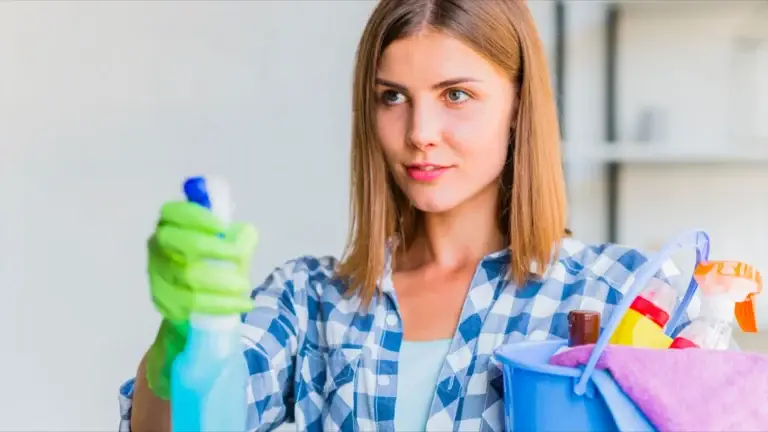 Woman in a blue plaid shirt and green rubber gloves holding a spray bottle and a blue bucket filled with cleaning supplies.