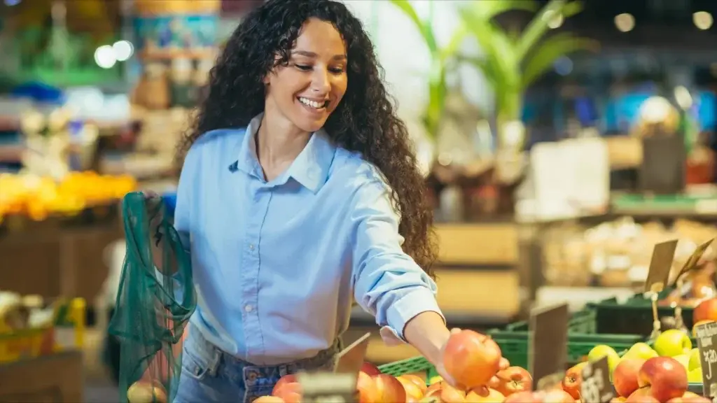 Smiling woman with reusable bag picking fresh fruit at a grocery store.