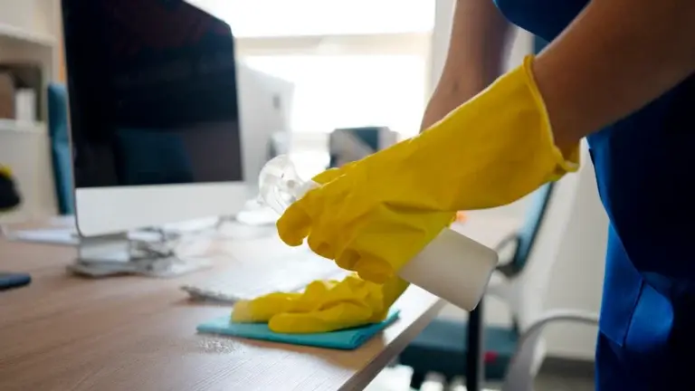 Cleaner in yellow gloves wiping an office desk with spray and a cloth.