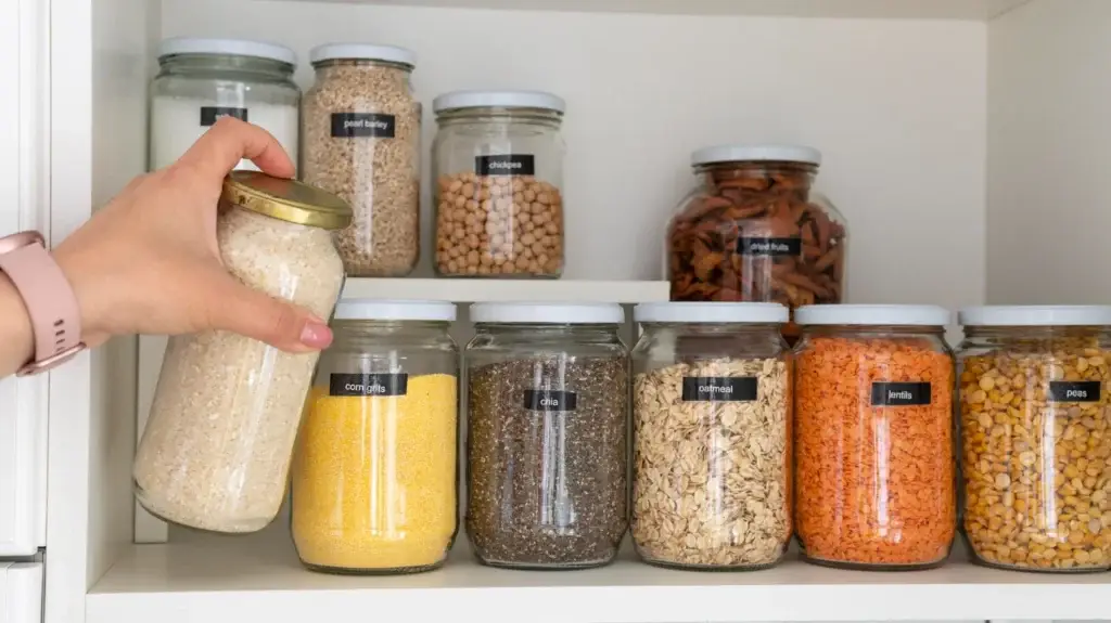 Hand placing a labeled glass jar among organized pantry containers on a white shelf.